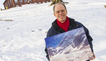 Peter Hansen stands in front of a snow-covered Boynton Hill, holding a painting of Mt. Everest. He's smiling, and is wearing glasses, a dark blue jacket, and red scarf.