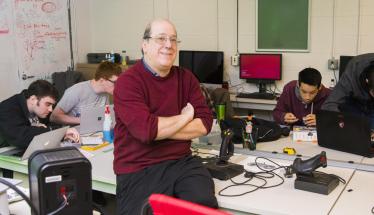Jeff Kesselman sits on the edge of a table with gaming equipment on it while students work in the background. He's smiling and is wearing dark pants, glasses, and a red sweater.