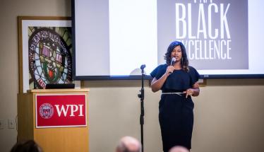 Whitney Gaskins stands near a WPI podium with a microphone in hand, addressing the audience. A projection screen reading "Black Excellence" is behind her, and she is wearing a black dress and silver belt.