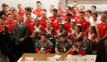 Keegan Concannon sits at a table with two friends, while the WPI football team and cheerleaders are gathered around him.