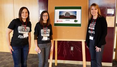 Three female Women in Data Science volunteers in matching T-shirts stand next to the official conference sign. They're all smiling and wearing lanyards, and one is wearing glasses.