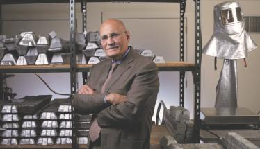 Diran Apelian stands in front of several shelves holding multiple pieces of metal. There's also a silver safety helmet behind him. He's got his arms crossed in front of him, and is wearing glasses and a dark suit and tie.