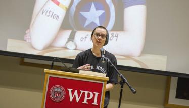 A WPI student stands at a podium as their Dear World portrait is displayed behind them, holding a piece of paper and sharing their story with the crowd.