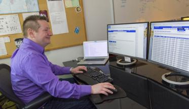 Craig Shue sits at a desk in front of three computer monitors. He's typing with one hand and mousing with the other. He's smiling, and is wearing a dark purple shirt, jeans, and silver watch.
