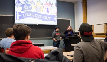 Students gather in a classroom to discuss a game, "Dragged Out," which is projected on screen while two students stand at a podium in mid-discussion.