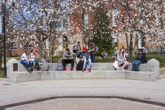 Several students sit on the concrete area around the Fountain in the center of campus as buds bloom on the trees behind them.