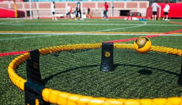 A close-up photo of a spikeball net with a ball bouncing off of it on Alumni Field during a sunny day.