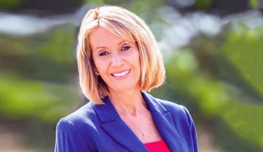 Marcia McNutt smiles in front of a natural background. The photo is from her shoulders up, and she is wearing a purple blazer and pink top.