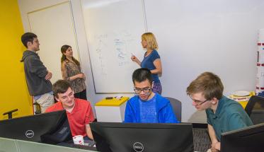 Two students stand at a white board in the background of the photo while their professor explains something to them. Three other students sit in the foreground, studying information on computers.