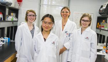Natalie Farny (center) and her students pose in their lab, between two lab tables. They're all smiling and are wearing lab safety coats and gear.