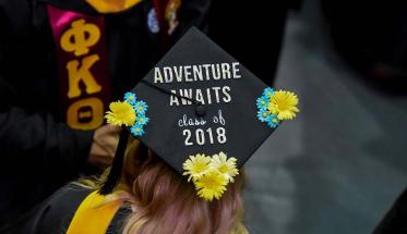 A close-up photo of a graduate's cap decorated with flowers and the phrase "Adventure Awaits: Class of 2018."
