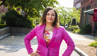 Bonnie Walker stands in the center of campus with her hands on her hips, the sunlight streaming in from behind her. She's wearing a pink blazer and white, pink, and orange patterned blouse.