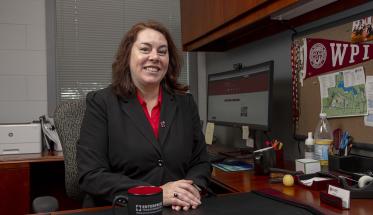Patty Patria sits at her desk in the IT office. She's smiling and is wearing a black blazer and red shirt with her computer and a WPI pennant in the background.
