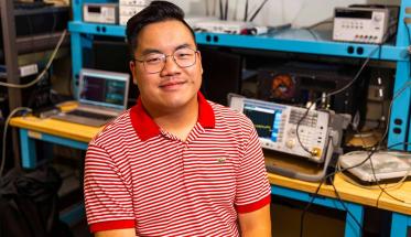 Max Li sits in a lab with a variety of tech equipment behind him. He's wearing glasses and a red and white striped shirt, and is smiling.