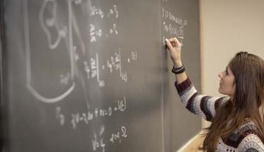 A woman writes a math equation on a chalkboard that already features many different equations and formulas.