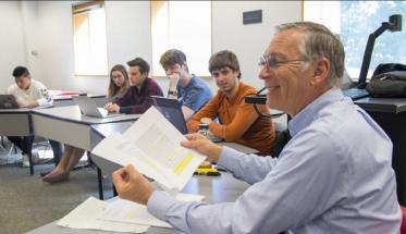 Craig Putnam sits in one of his classes with students' desks arranged in a circle. He's smiling and is holding several papers in his hands.