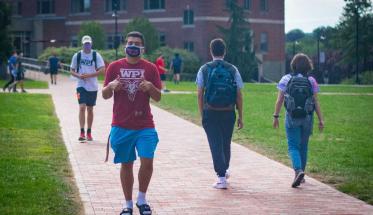 Students walk through the Quad wearing face coverings and maintaining social distance. One flashes two thumbs up at the camera.