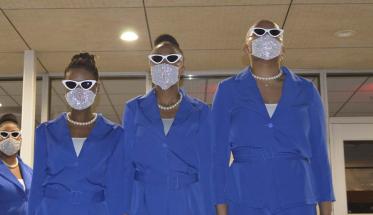 Four new members of Zeta Phi Beta stand in front of Harrington Auditorium wearing matching outfits, masks, and sunglasses.