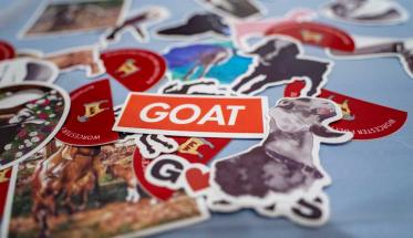 A pile of stickers, one of which says GOAT in white letters against a red-orange background.