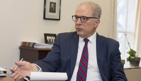 Bogdan Vernescu, wearing brown glasses, a dark blue suit jacket, white button-down shirt, and red and white striped tie, sits at a desk and is in mid-discussion.