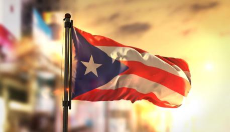 A photo of the Puerto Rican flag with yellow and orange sunshine in the background, looking as if it was taken at sunset or sunrise.