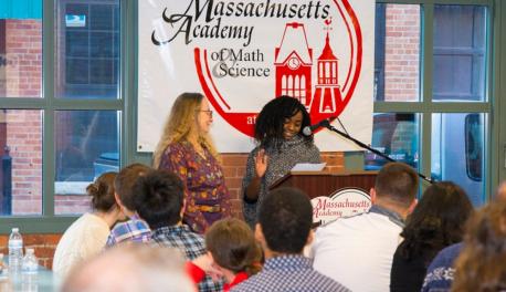 A student stands at the podium addressing a former teacher. They're in front of a brick wall and a banner with the Mass Academy logo, and two large windows.