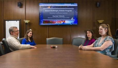 Two faculty members sit across a conference table from two students, with a computer screen projected in between them that reads, "Grand Challenges Scholar Program of Worcester Polytechnic Institute."
