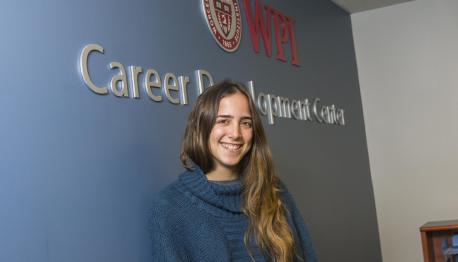 Camila Di Fino Napolitano stands in front of a blue wall with "WPI Career Development Center" written on it. She has long brown hair and is smiling and wearing a dark blue sweater.