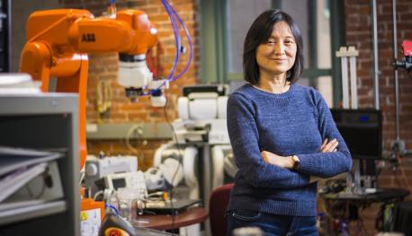 Jing Xiao stands in a robotics lab with a brick wall and several different machines and robots behind her. She has black hair, is smiling, has her arms crossed, and is wearing a blue sweater and jeans.