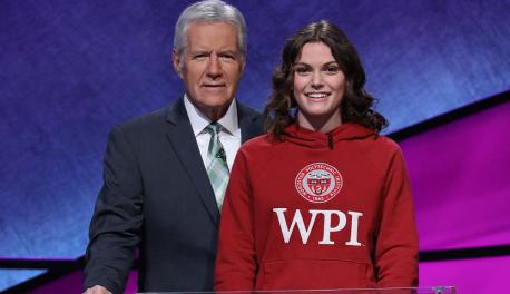 Alex Trebek (left) is wearing a suit and standing next to Alli Ross, who is wearing a red WPI sweatshirt, on the Jeopardy! set. Both are smiling.