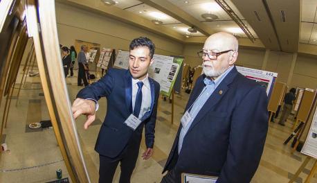 A student dressed in a professional suit and tie gestures toward his poster presentation while explaining a concept to a judge.