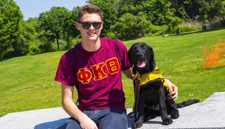 Jake Scheide sits in Institute Park with Diesel by his side. He's smiling and wearing a red fraternity shirt and sunglasses. Diesel's tongue is out, he has black fur, and is wearing his yellow training cape.