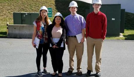 Four students wearing white hard hats and protective glasses and three carrying clipboards smile together at their project site in Boston, Mass.