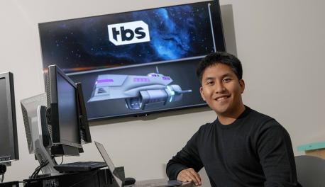 Joshua Galang smiles in front of a projection screen showing a model of an animation from Final Space, as well as the Final Space logo, with his laptop in front of him.