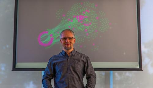 Dmitry Korkin stands before a depiction of a network of proteins that are affected by type 2 diabetes (in pink). The lines represent protein-protein interactions that are expected to affected by the mutations that are linked to types 2 diabetes.