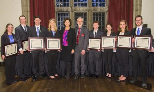 Two teams of four students each stand on either side of President Leshin and Kent Rismiller. All are dressed in professional attire, the students are holding framed certificates, and there are large windows with red curtains in the background.