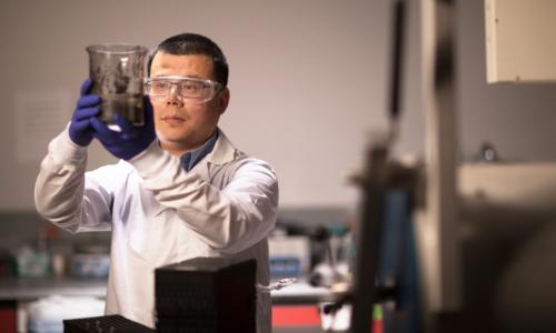 Yan Wang, dressed in a lab coat, holds up and looks at a large beaker containing a black material