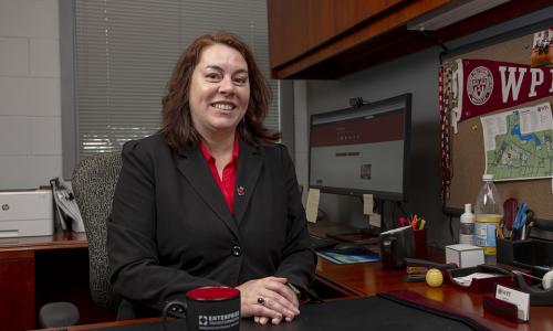 Patty Patria sits at her desk in the IT office. She's smiling and is wearing a black blazer and red shirt with her computer and a WPI pennant in the background.