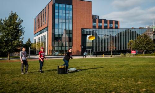Students play frisbee on the Quad with the Innovation Studio in the background.