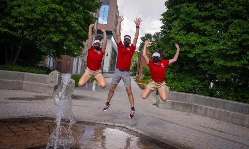 Three orientation leaders jump in unison behind the fountain.