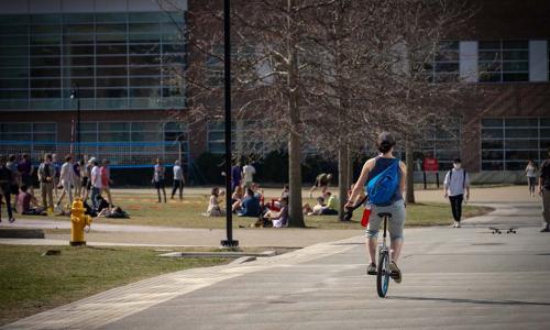 A student rides a unicycle past a busy quad.